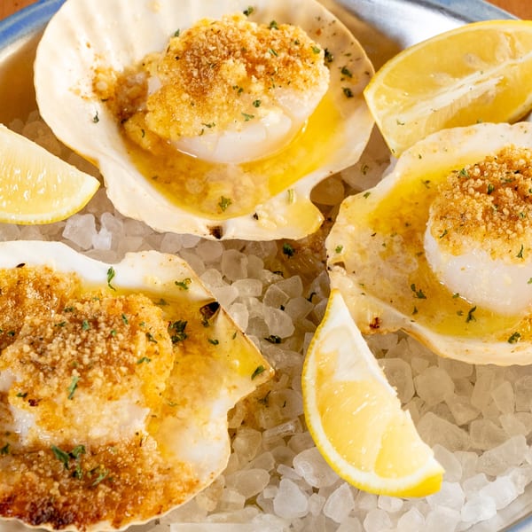 Three baked scallops in shells with breadcrumbs, butter, and lemon wedges on a bed of ice.