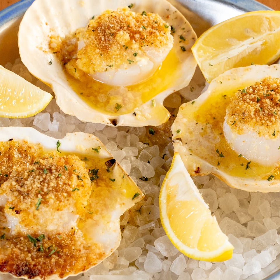 Three baked scallops in shells with breadcrumbs, butter, and lemon wedges on a bed of ice.